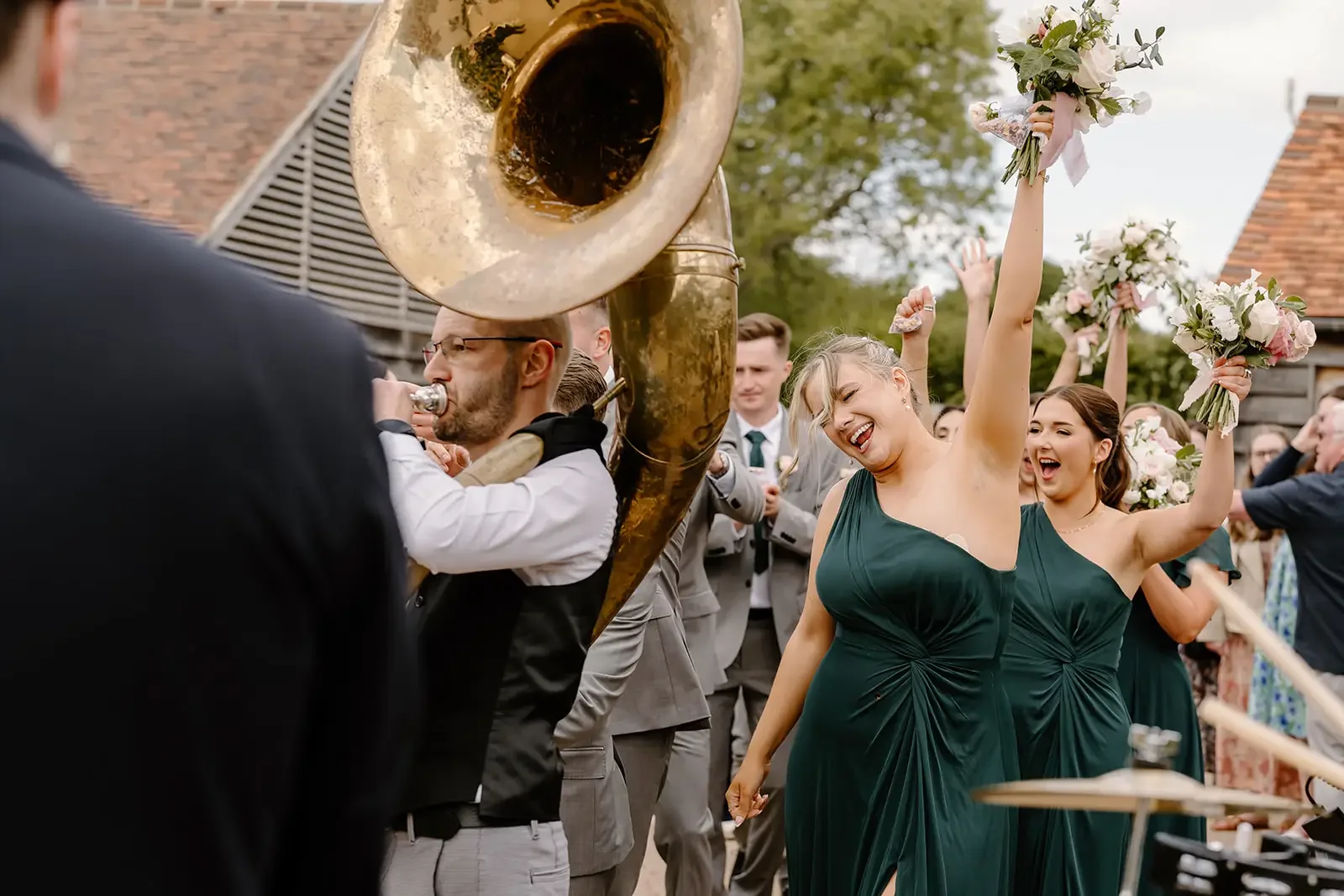 Group of people at a wedding celebration, with women holding bouquets and raising their arms in joy, and a man playing a sousaphone in the foreground.
