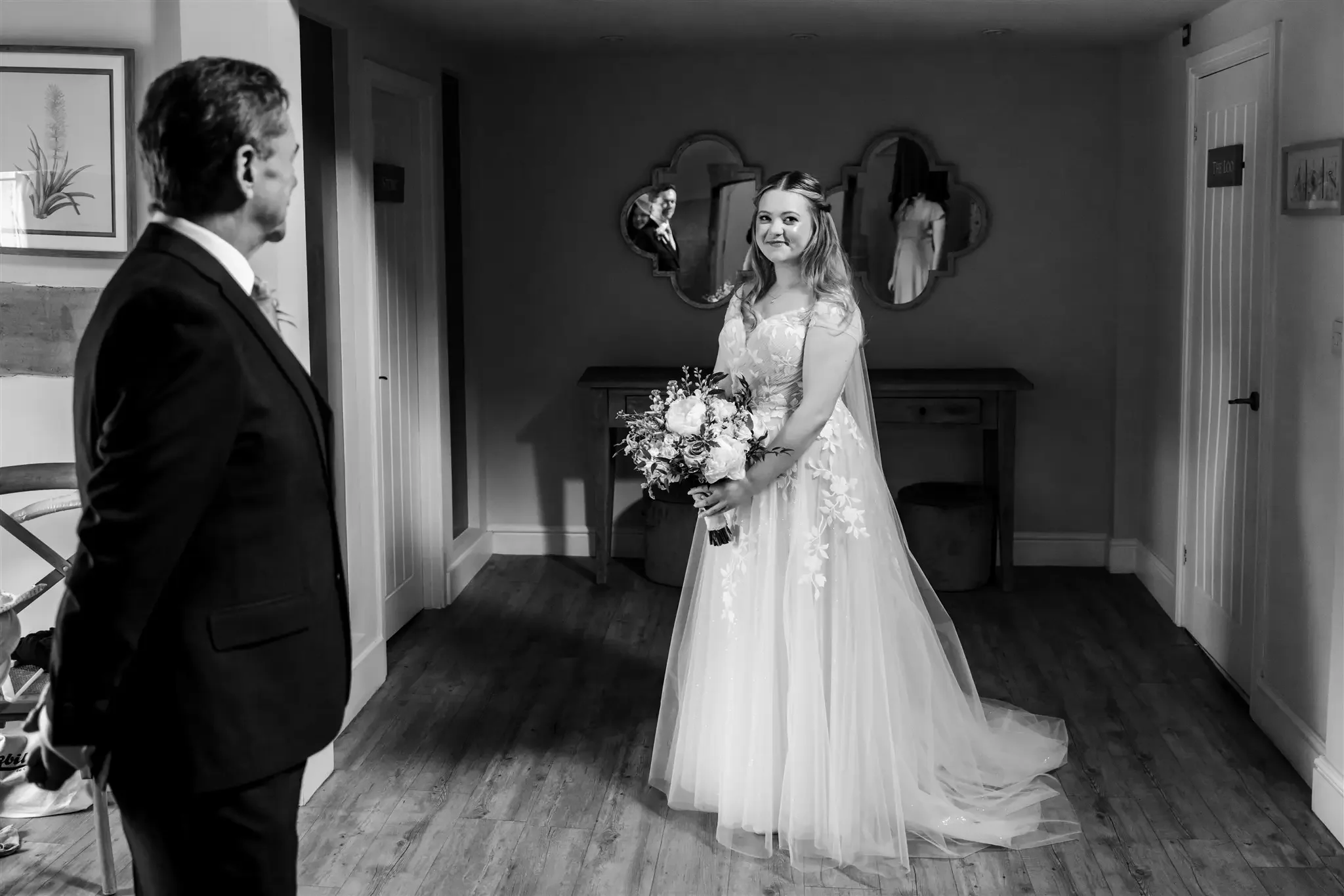 A bride in a wedding dress holding a bouquet looking at a man in a suit in a room with mirrors on the wall.