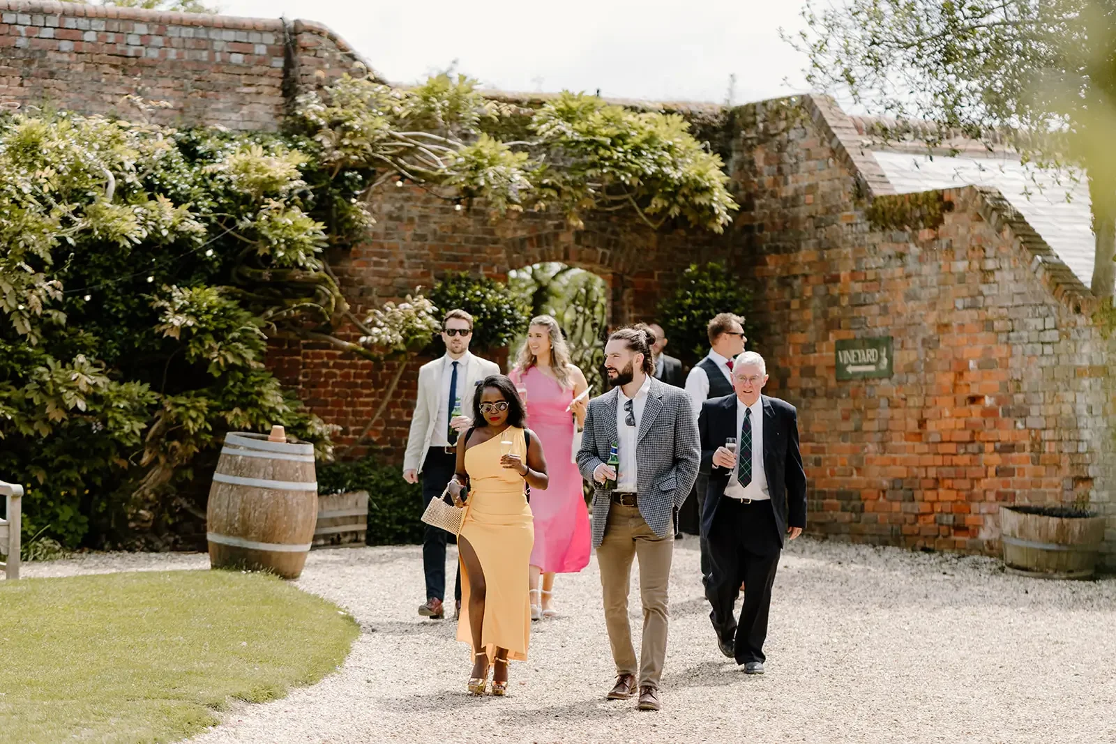 Group of well-dressed people walking outdoors near a brick wall with greenery during daytime, some holding drinks.