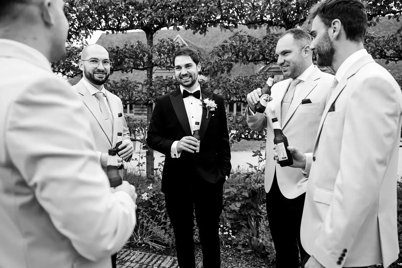 A group of five men dressed in tuxedos and suits, smiling and holding bottles of beer, standing outdoors in a garden or park with trees and plants in the background.