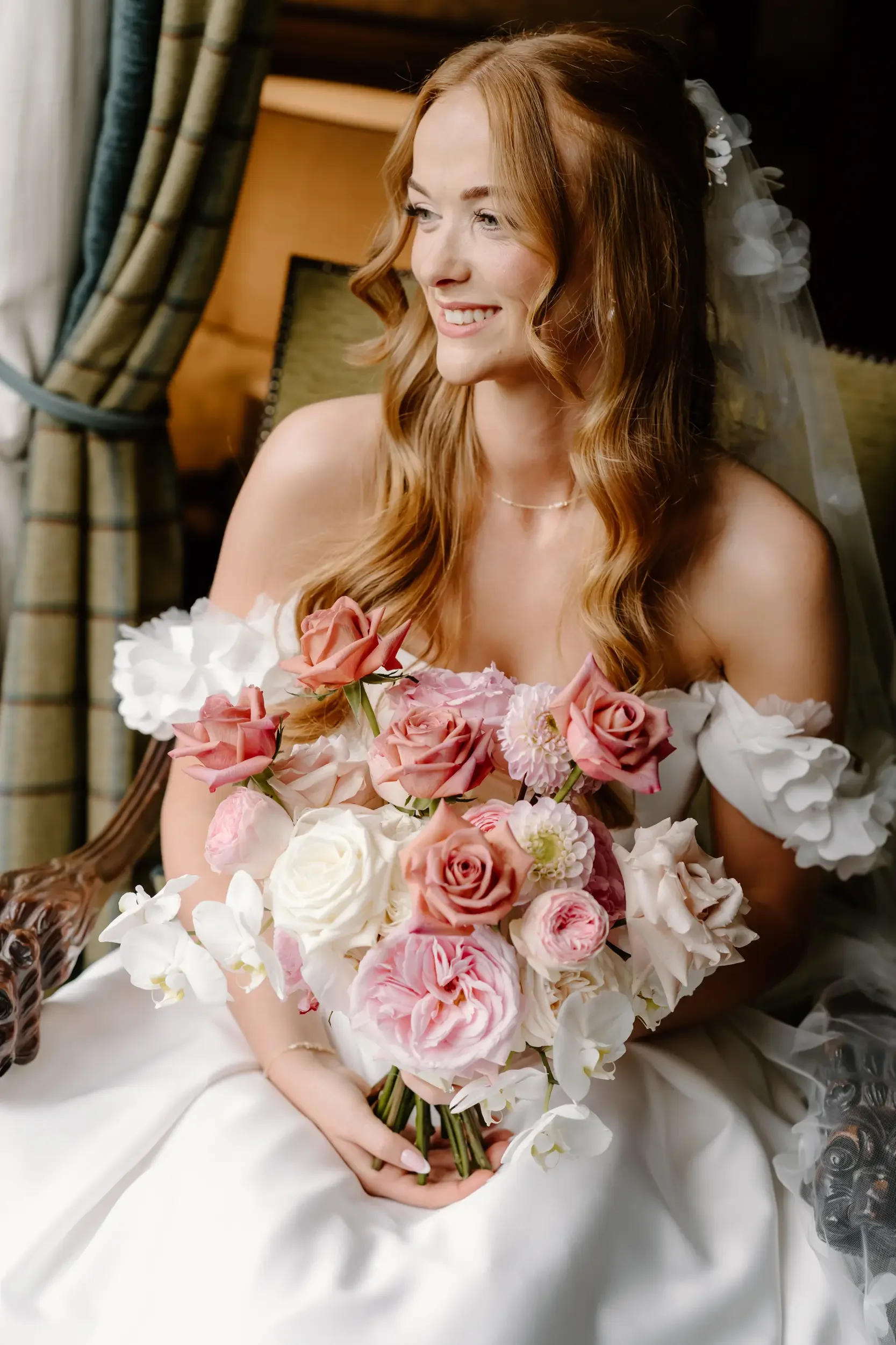A smiling bride with red hair holding a bouquet of pink, white, and blush roses and orchids, wearing a white wedding dress with ruffled off-shoulder sleeves, sitting indoors near curtains.