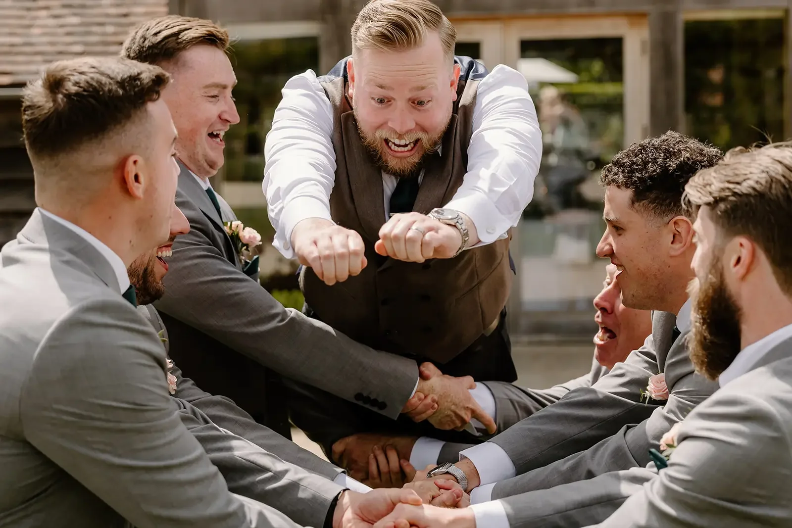 A man leaping over a table of men in suits during a wedding celebration, everyone is smiling and laughing.