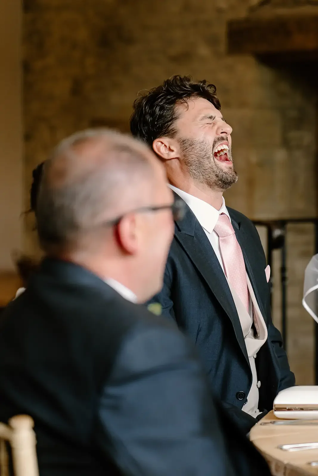 A man in a suit and pink tie laughing at a formal event, with another man in glasses in the foreground.