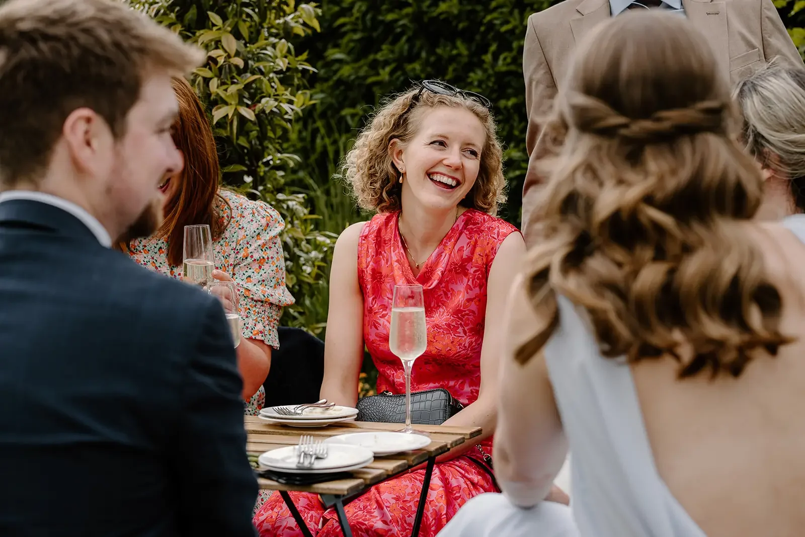 A group of people having a conversation outdoors, with a woman in a red dress smiling and holding a glass of champagne, surrounded by lush green bushes.
