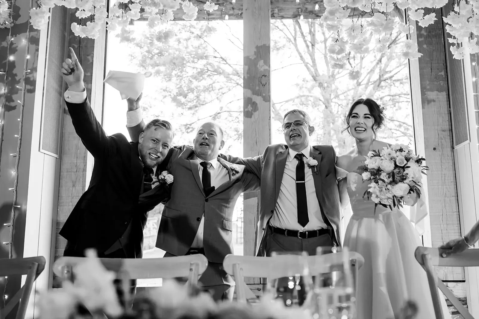 Black and white photo of a wedding reception, with four people smiling and standing in front of a large window with trees outside. Two men are in suits, one holds a tissue, and a woman in a wedding dress holds a bouquet of flowers.
