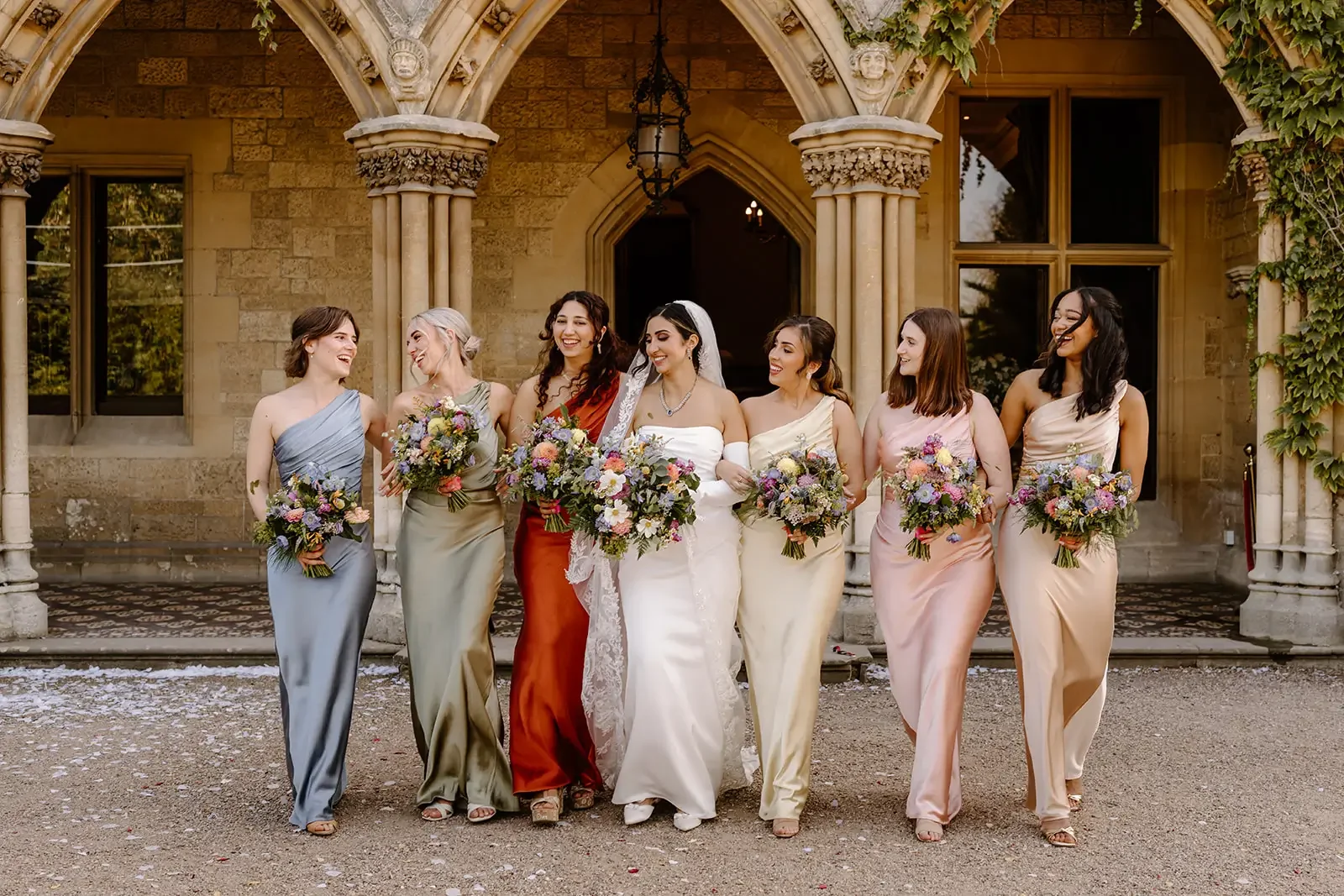 A bride and her six bridesmaids walking together in front of a stone building with arched entrance, all holding bouquets of flowers and smiling.