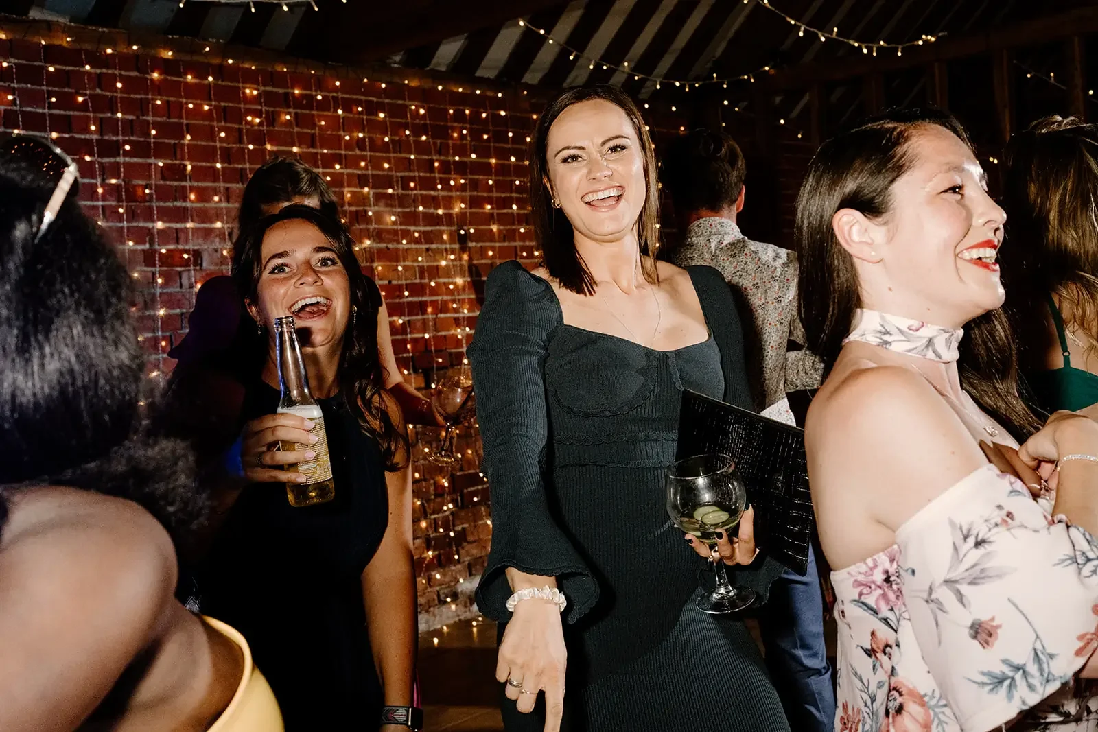 People enjoying a party or celebration indoors with string lights and a brick wall in the background. Several women are smiling and holding drinks, dressed in stylish outfits.