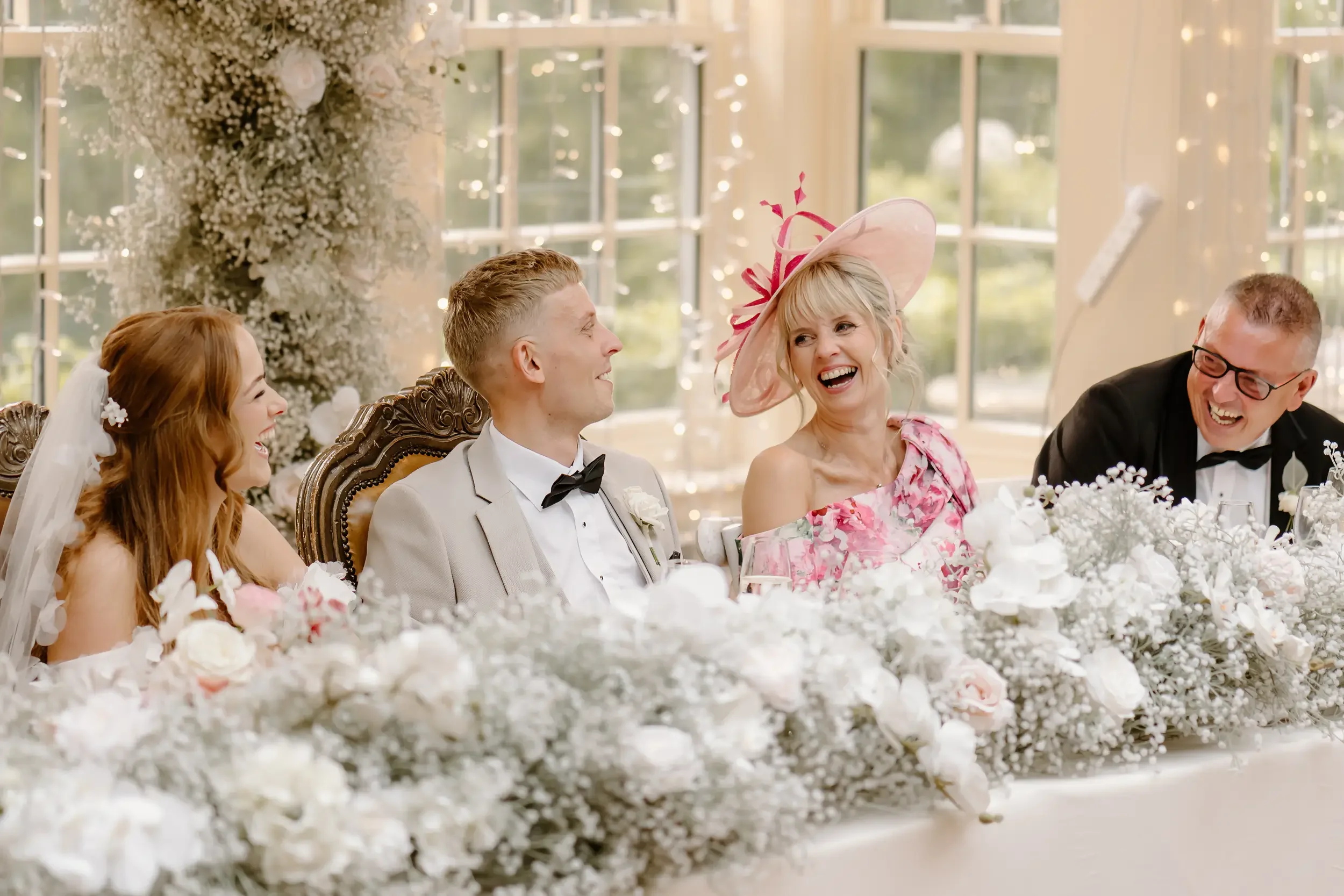 People sitting at a wedding reception, laughing and smiling, with a decorated floral table and large windows in the background.