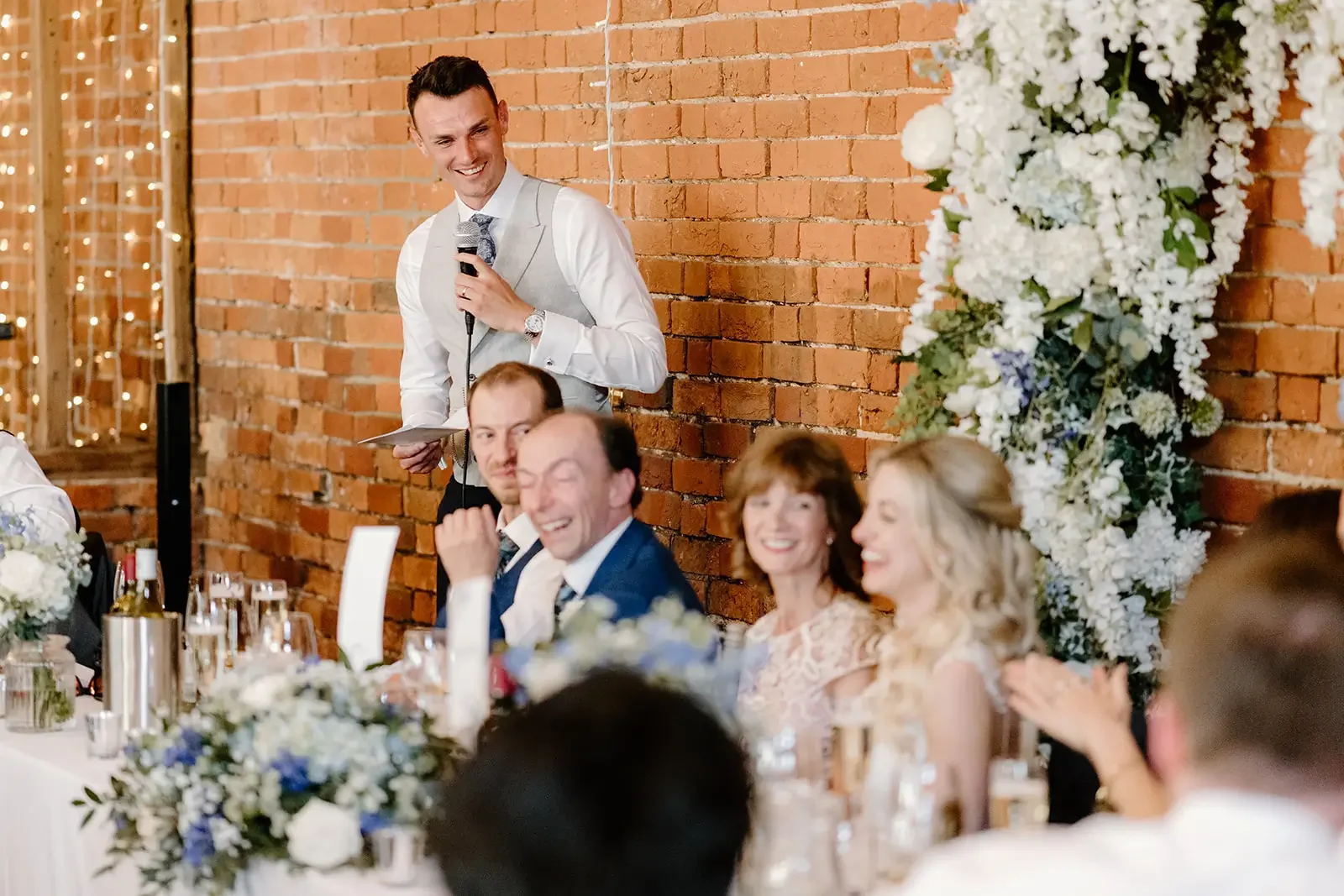 A man giving a speech at a wedding reception, standing behind a table with seated guests, with an exposed brick wall and floral decorations in the background.