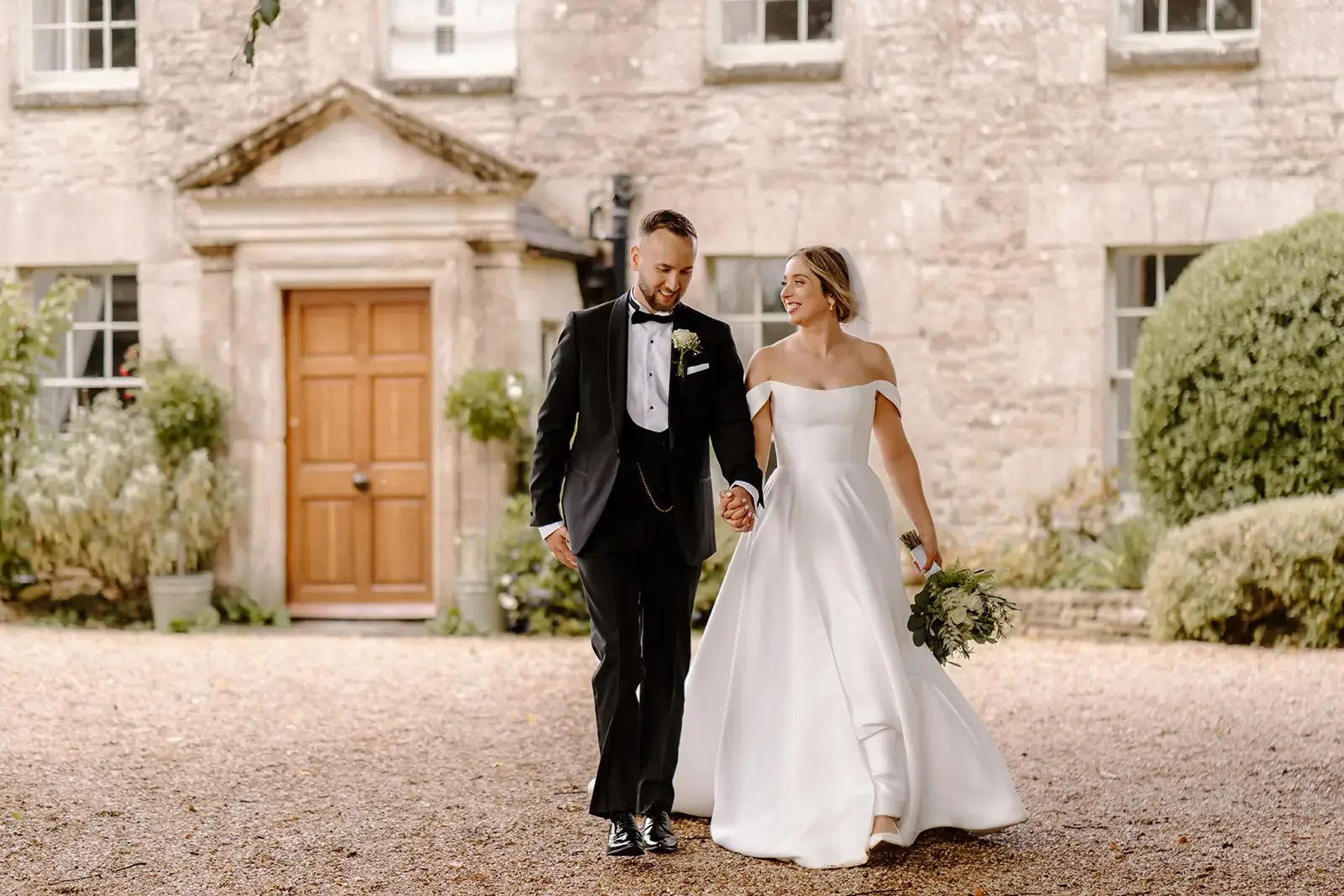 Bride and groom walking hand in hand outside a stone building, wedding attire, she carries a bouquet, smiling and looking at each other.