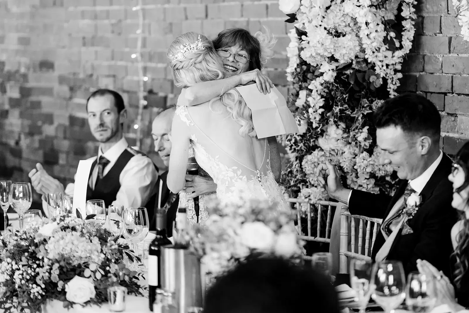 Two women embracing at a wedding reception, one with long hair and a flower crown, the other with glasses and short hair, while seated at a table with wedding guests, flowers, and drinks.