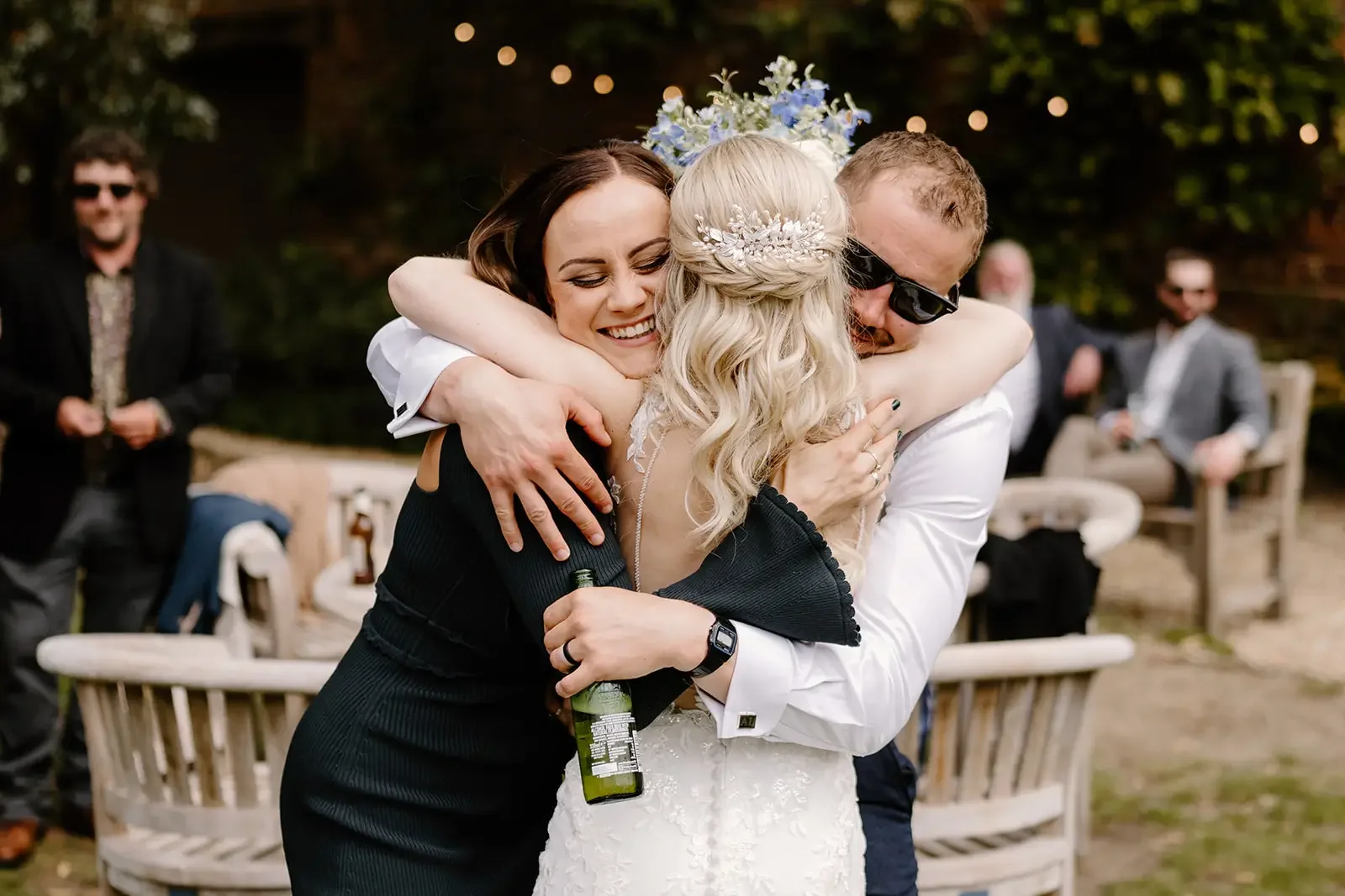 People hugging at outdoor wedding reception, woman in wedding dress with floral headpiece, man with sunglasses, woman in black dress, smiling and celebrating.