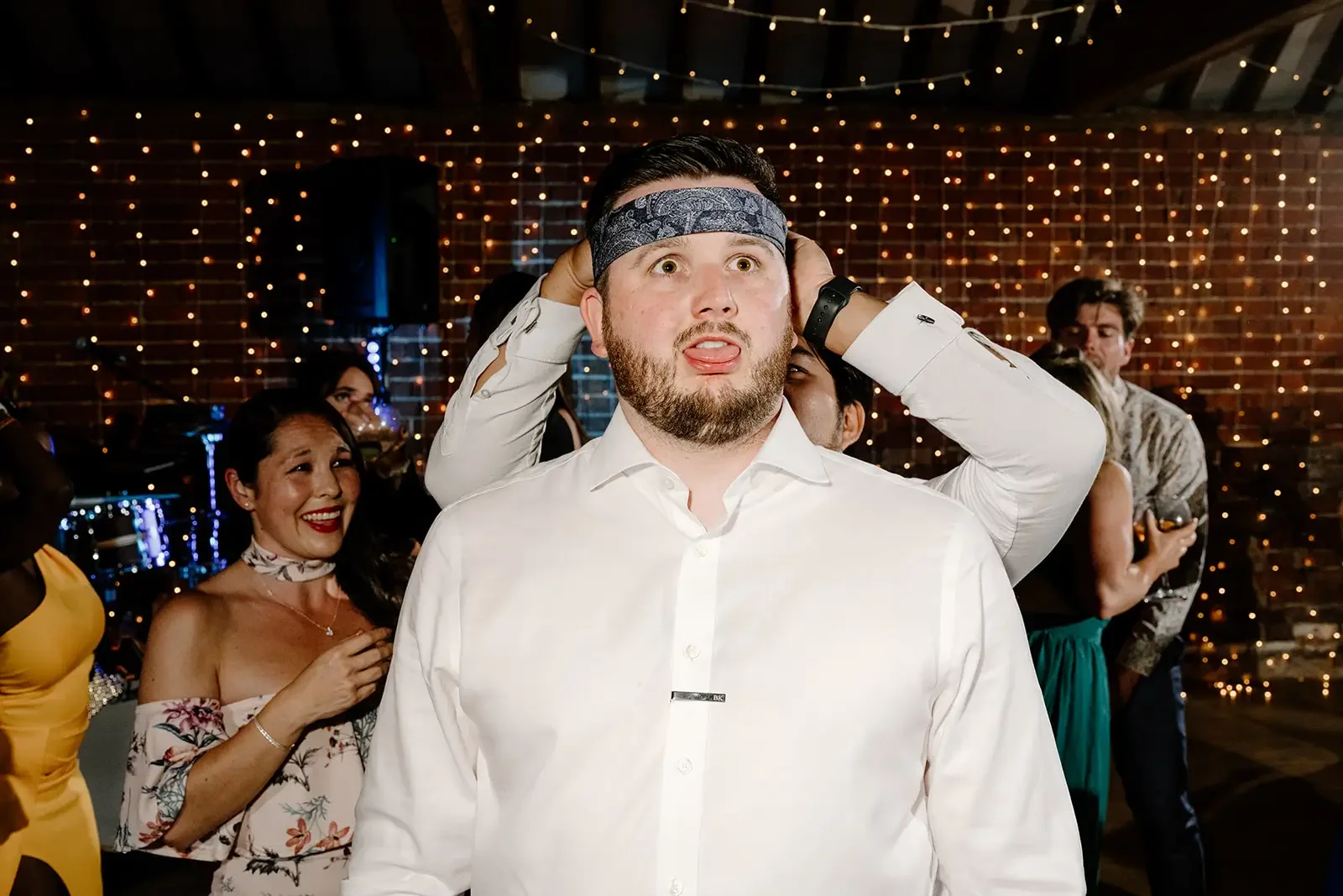 A man with a beard and white shirt placing a bandana on his head at a party or celebration with people and string lights in the background.