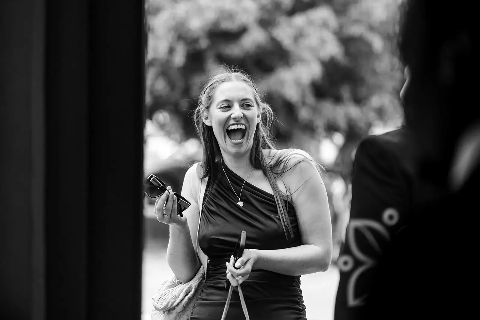 A woman with long hair smiling and laughing, holding sunglasses and a cellphone, engaged in a lively conversation outdoors.