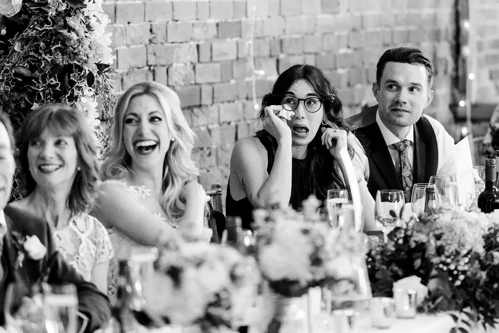 People sitting at a table during a celebration, with a woman in glasses wiping her eye and three others laughing and smiling, against a brick wall background.
