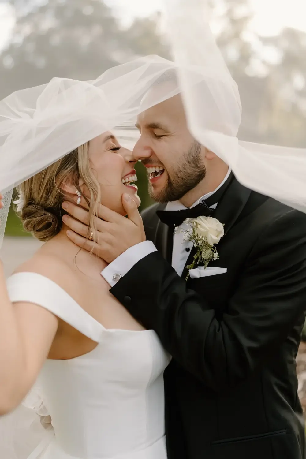 A bride and groom share a joyful moment under a veil on their wedding day, with the bride smiling and the groom touching her face, both dressed in formal wedding attire.