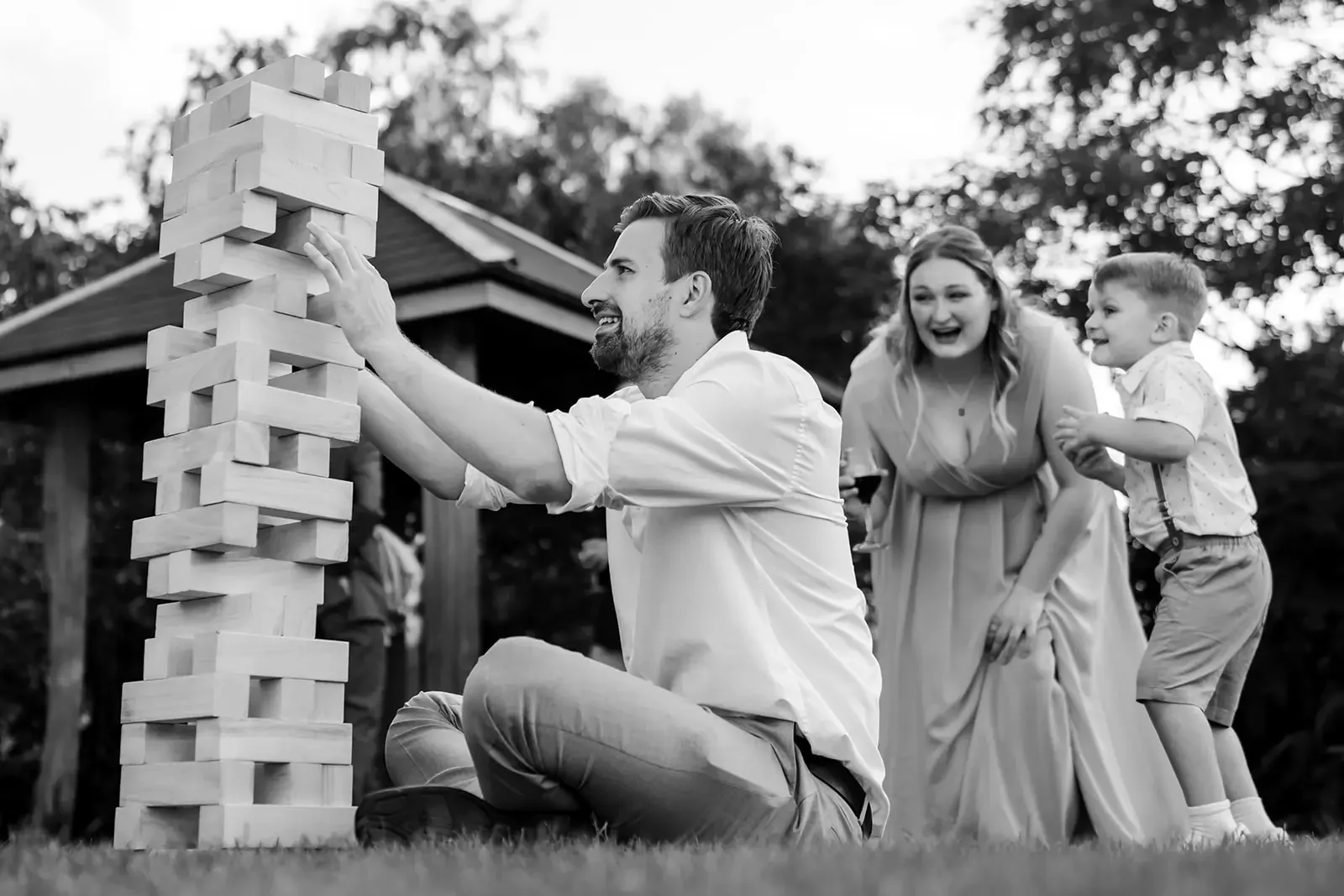 A man kneeling on the grass, playing a giant outdoor Jenga game with a woman and a boy, all smiling and laughing, in a park-like setting.