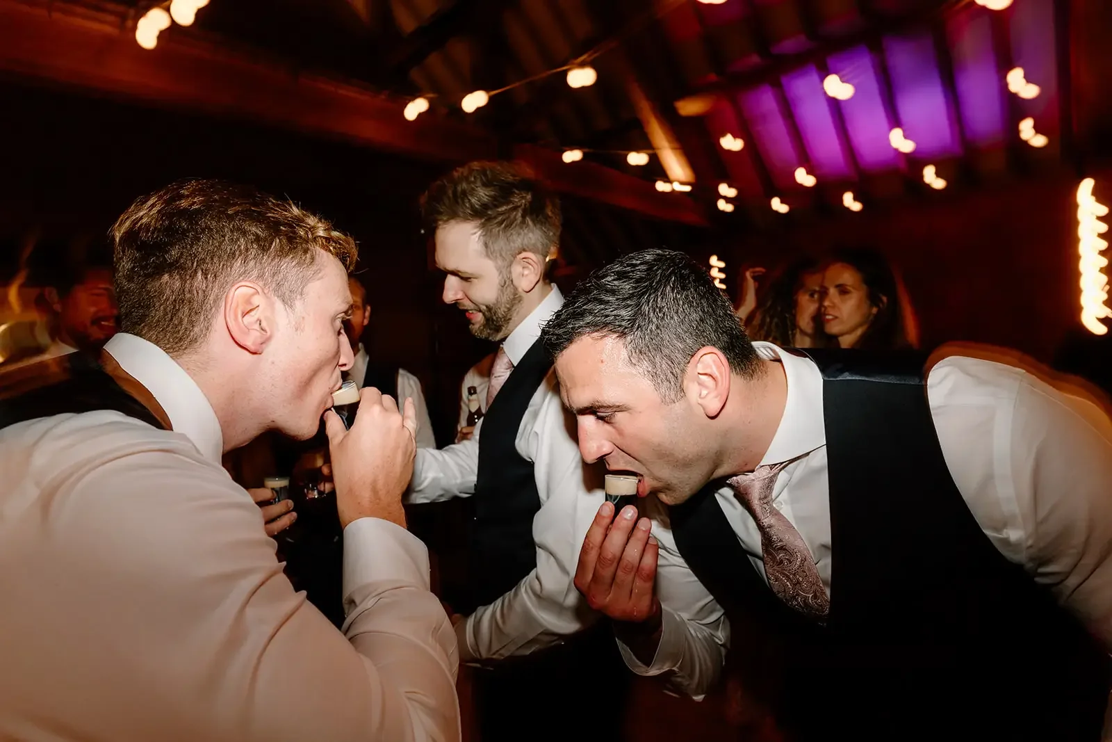 Men in formal attire enjoying shots at a celebration or party in a dimly lit setting with string lights overhead.