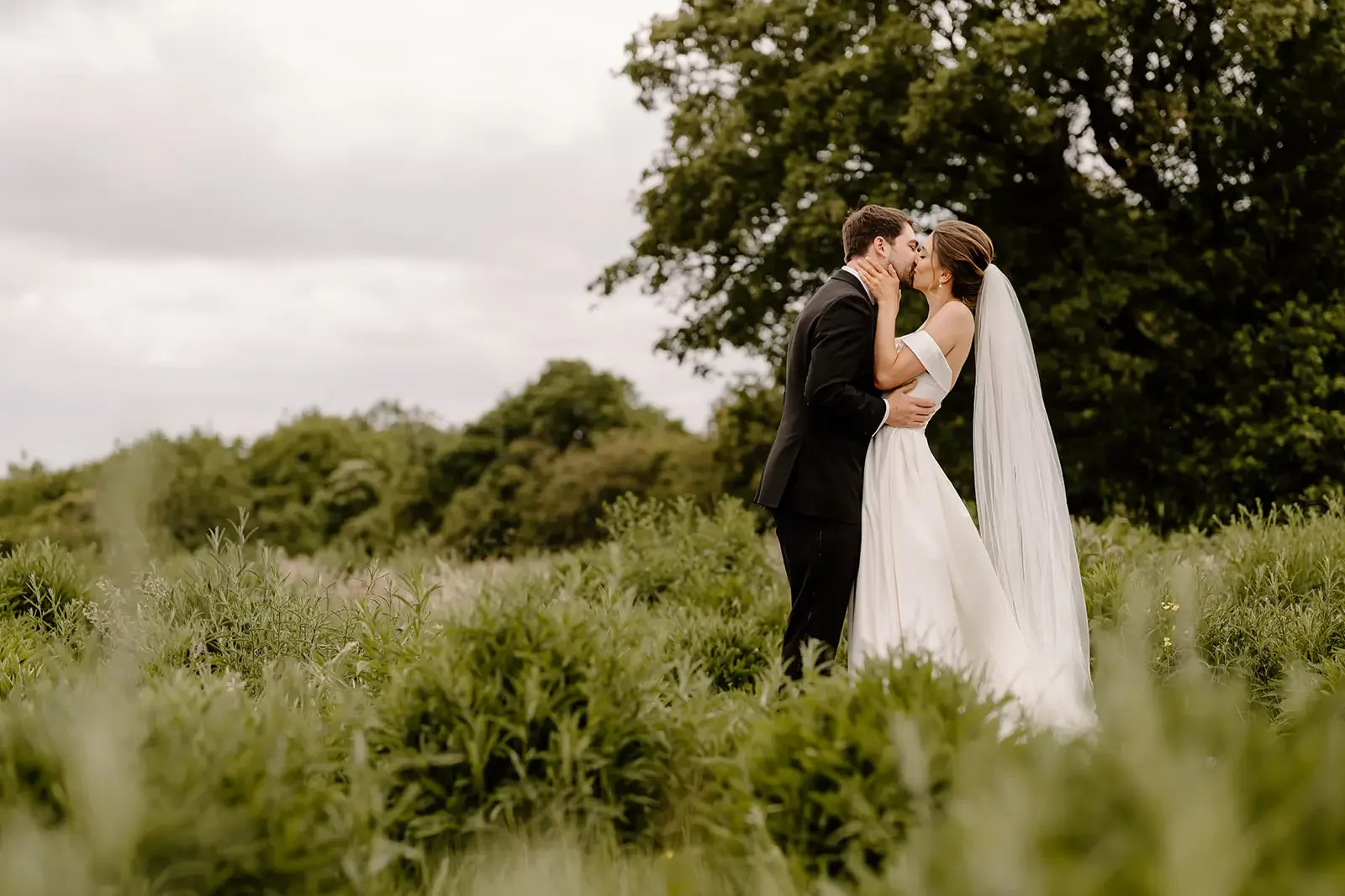 A bride and groom kissing outdoors in a green field with trees in the background.