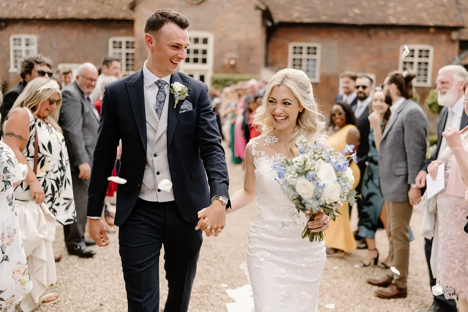 Bride and groom smiling and holding hands during their wedding, surrounded by family and friends outdoors.