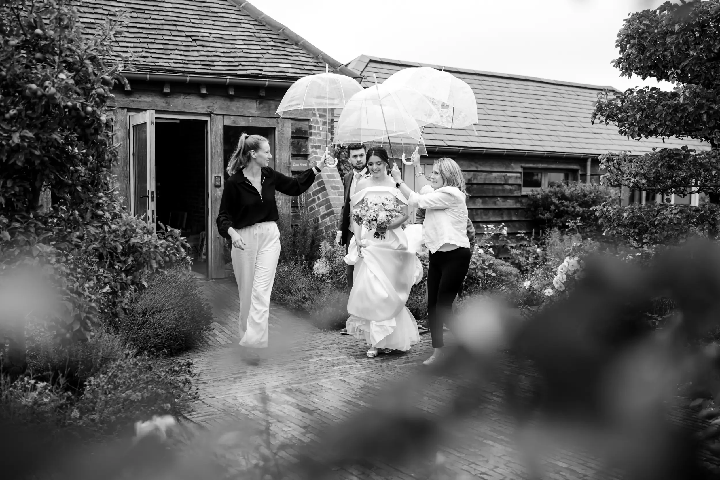 Black and white photo of a bride in a wedding dress holding a bouquet, walking outdoors on a brick path, assisted by two women, with two men following behind. Several people are holding transparent umbrellas over the bride and the group, amid a garde