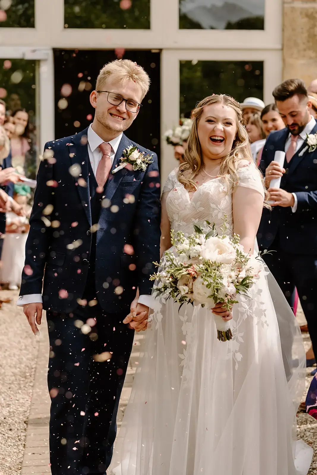 A bride and groom holding hands and smiling as they walk outside, surrounded by wedding guests, with confetti falling around them.