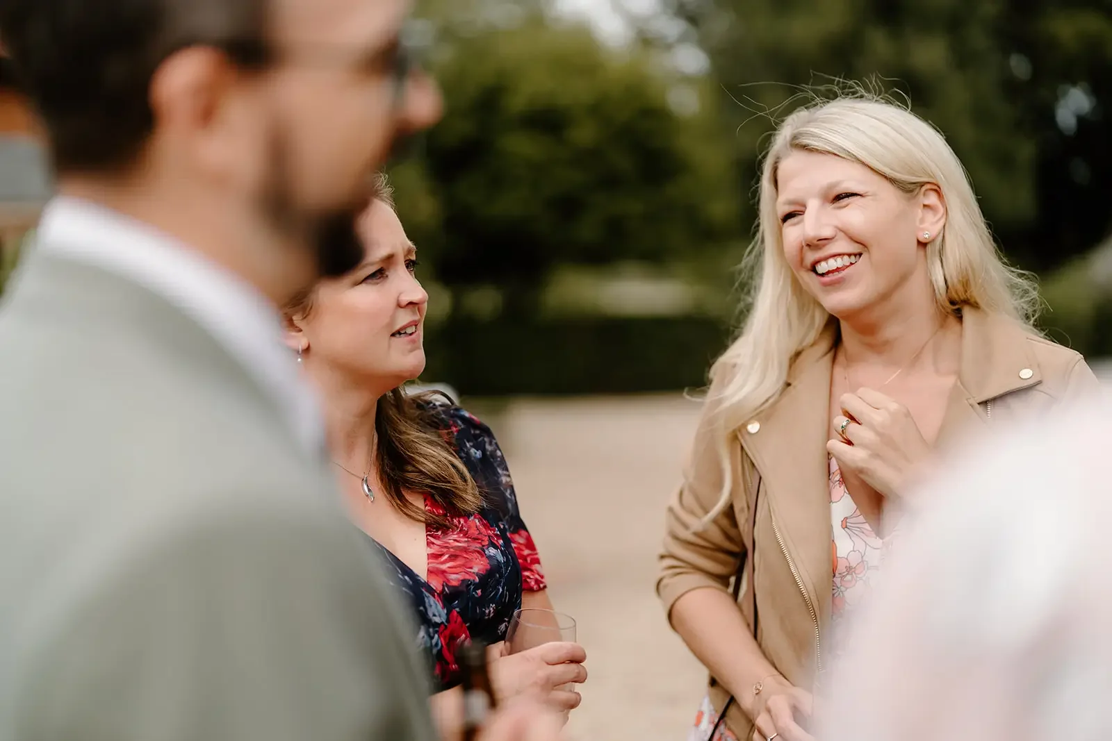 People socializing outdoors, four women and one man, with trees in the background, having a conversation and holding drinks.