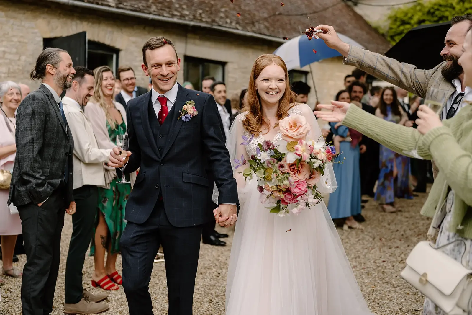 Newlywed couple holding hands and smiling, surrounded by friends and family celebrating outside during a wedding.