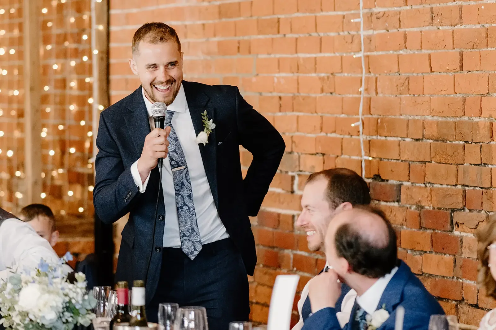 A man giving a speech at a wedding reception, holding a microphone, dressed in a dark suit with a white shirt and patterned tie, standing near a brick wall, smiling, with guests seated at a decorated table.