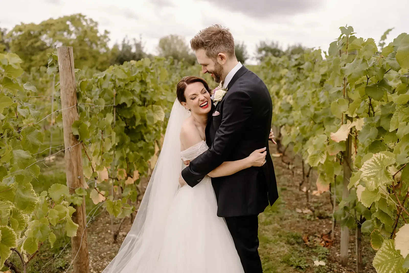 A bride and groom hugging and smiling in a vineyard, with lush green grapevines on either side.