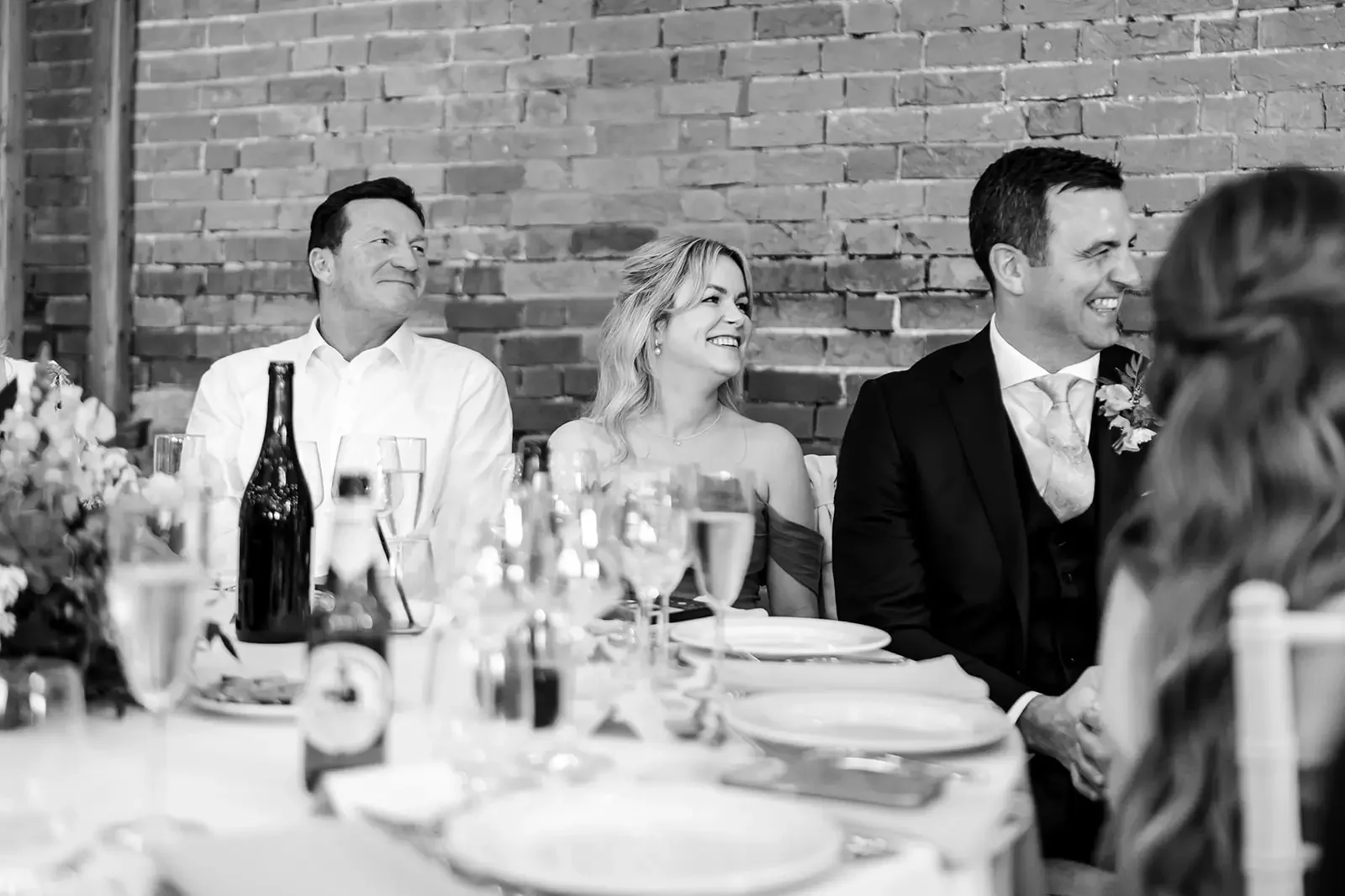 Black and white photo of people sitting at a table during a celebration, with a brick wall in the background. The people are smiling and appear to be enjoying the event.