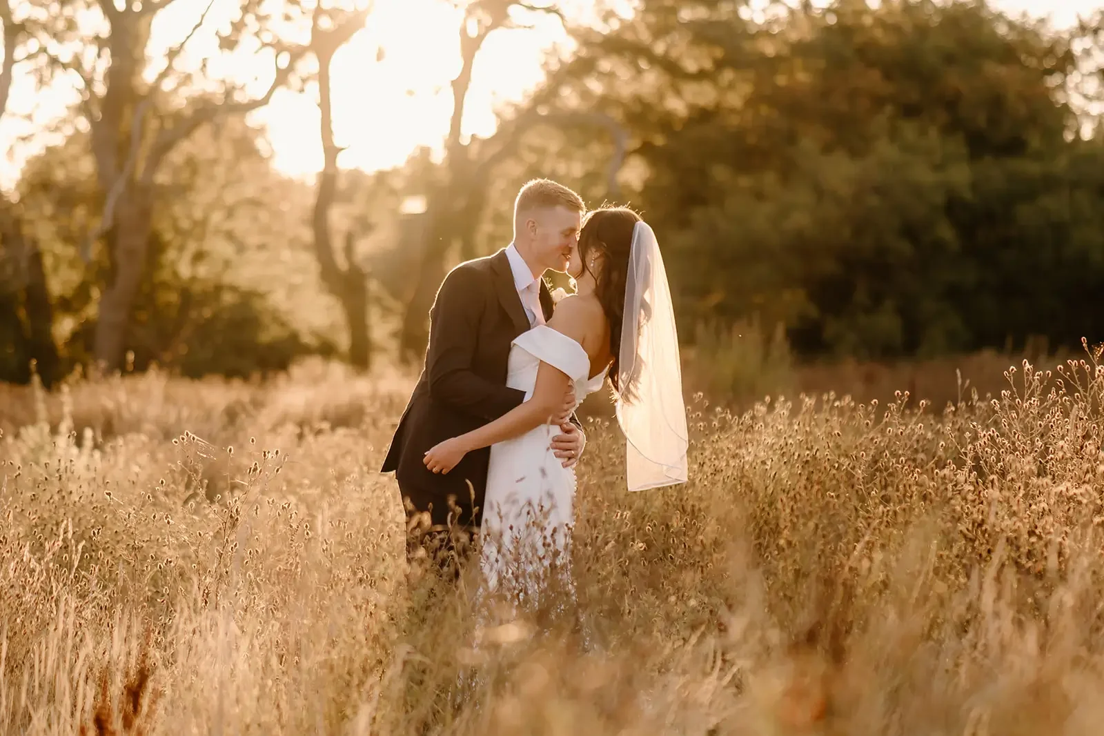 A couple in wedding attire sharing a kiss in a field of tall grass at sunset, with trees in the background.