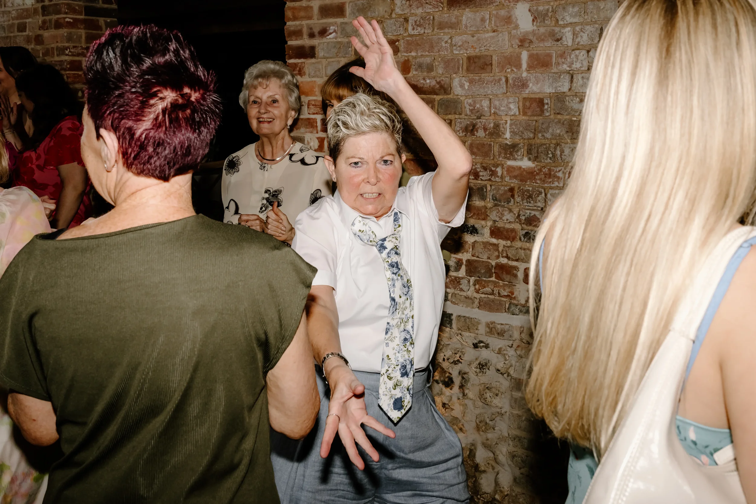 A woman with gray hair, wearing a white blouse and a patterned tie, appears to be dancing or throwing her hands up at a social gathering among mostly women standing close together against a brick wall.