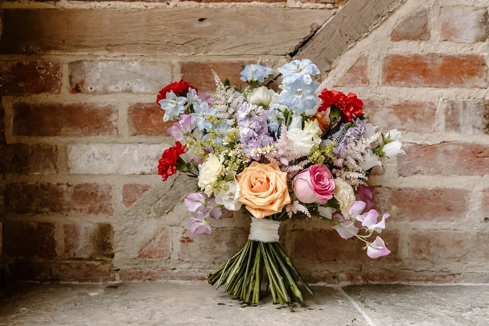 A colorful bouquet of various flowers including roses, carnations, and blossoms, tied with a white ribbon, against a rustic brick and wood background.