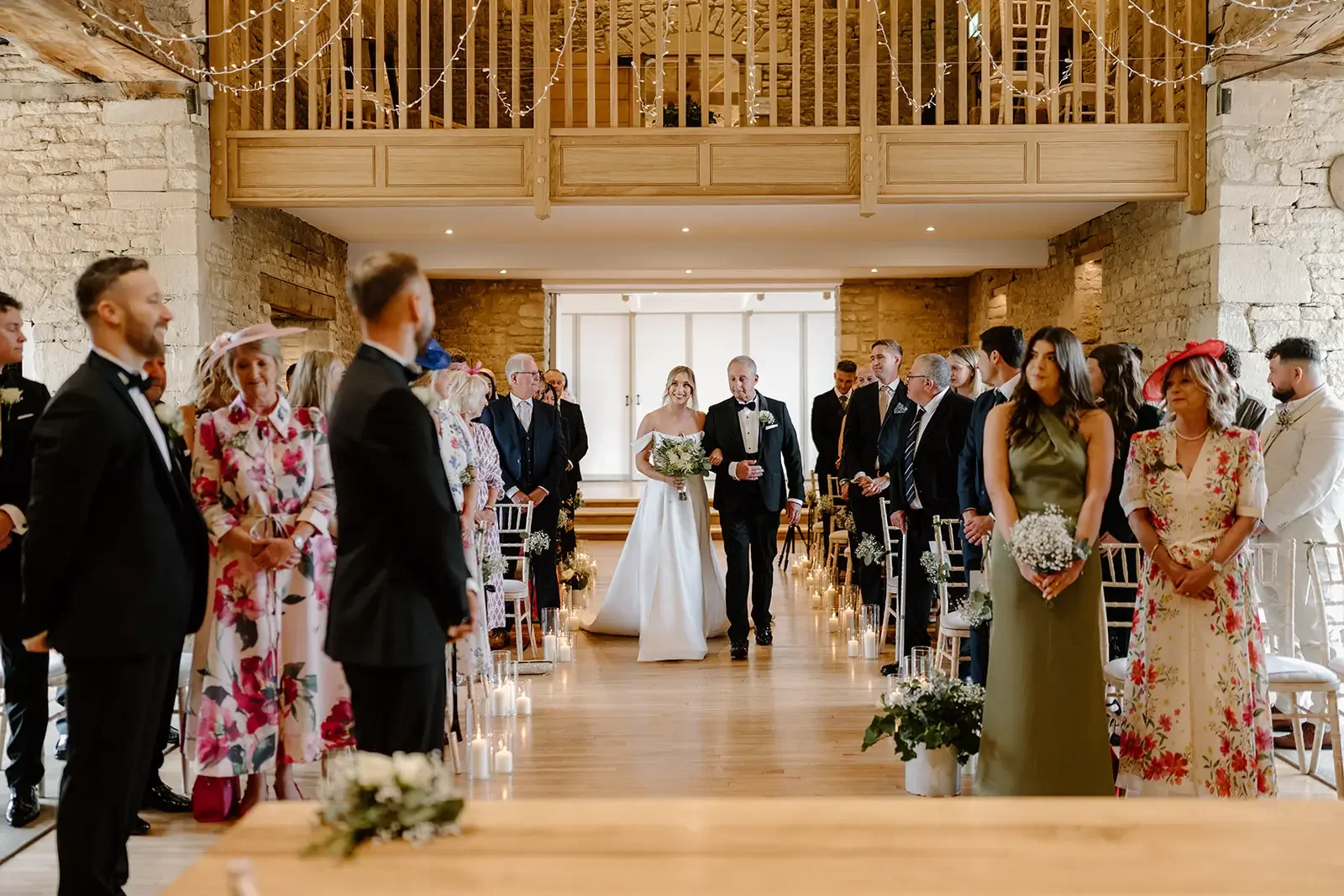 A bride walking down the aisle with her father at a wedding ceremony in a rustic venue with stone walls and wooden accents, surrounded by guests.