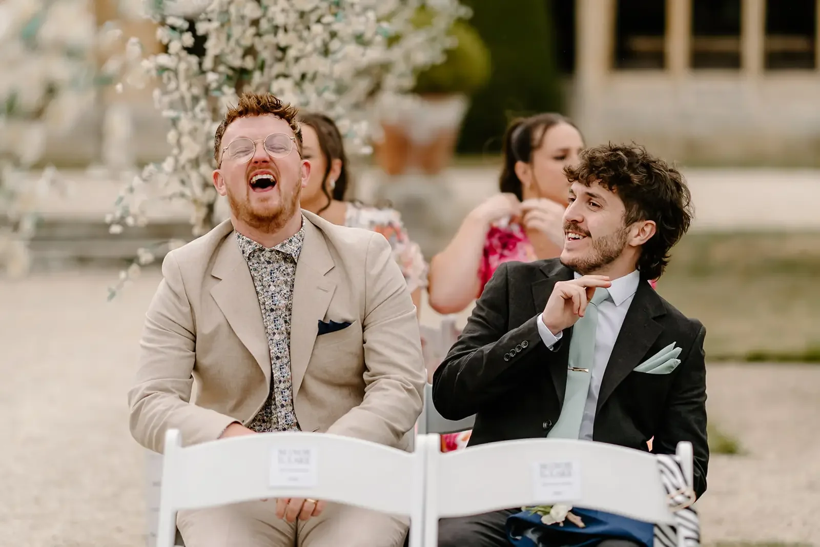 Two men sitting outdoors at a wedding or event, laughing and smiling, with women in the background.