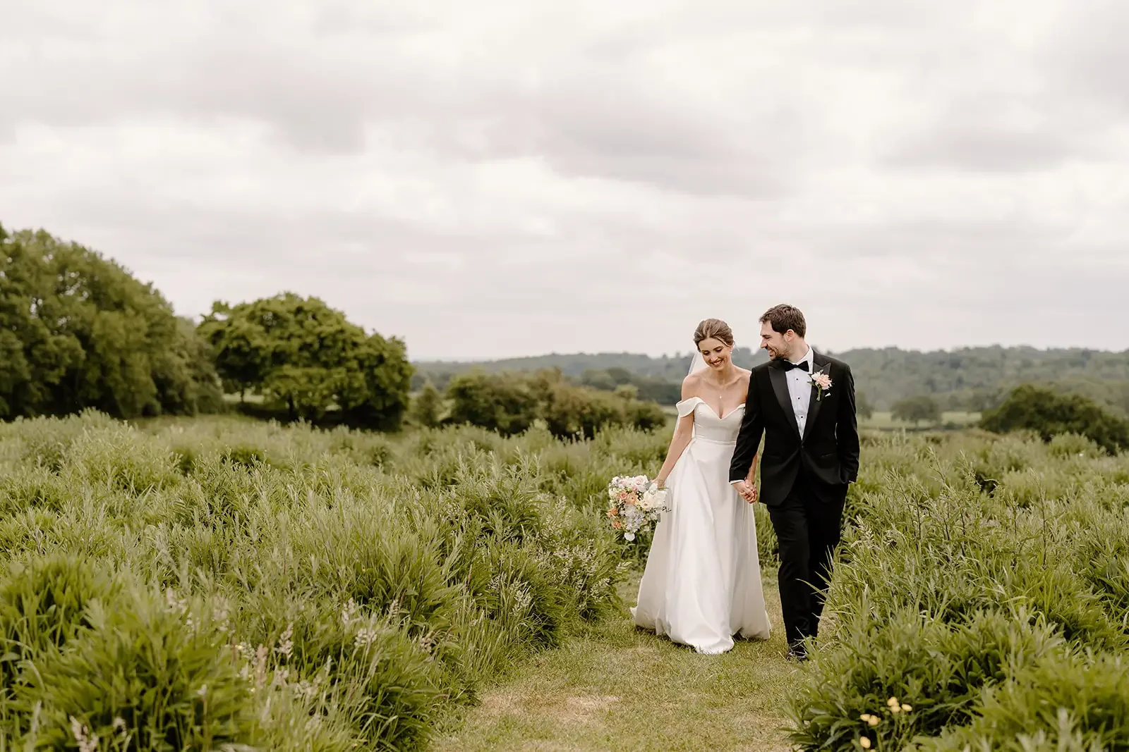 Bride and groom walking hand in hand through a green field on their wedding day, with the bride holding a bouquet of flowers and both smiling.
