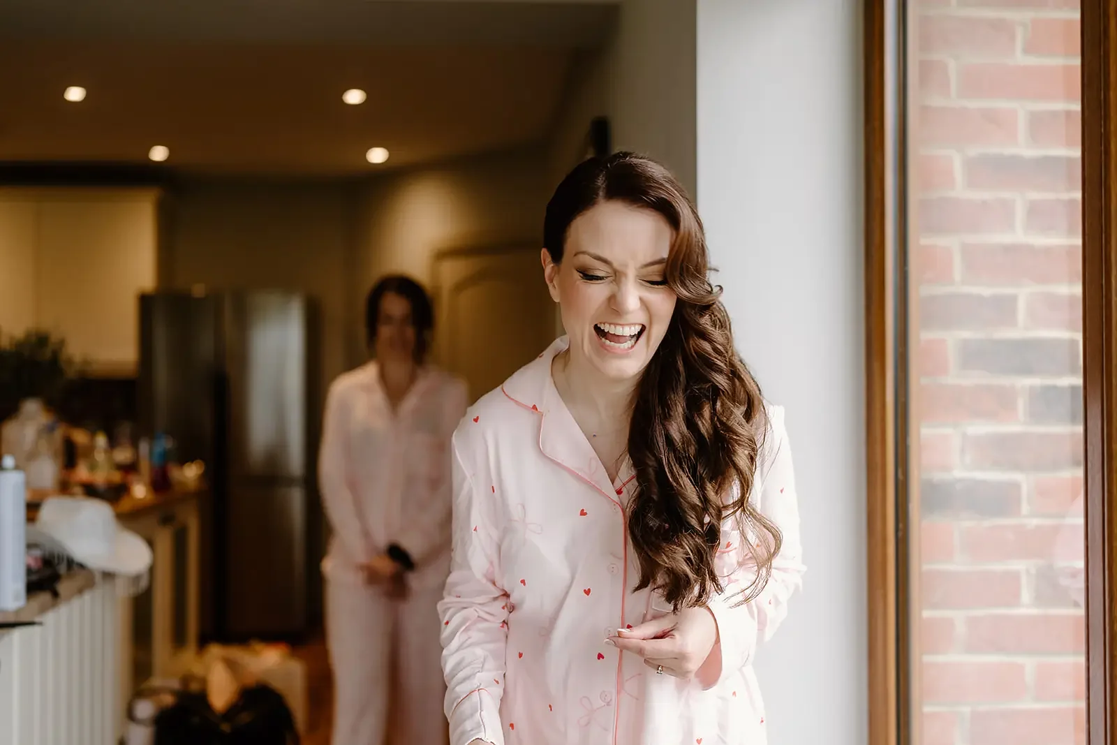 A woman with long curly brown hair laughing and wearing pink pajama set with small red hearts, standing by a window, with another woman in similar pajamas in the background.