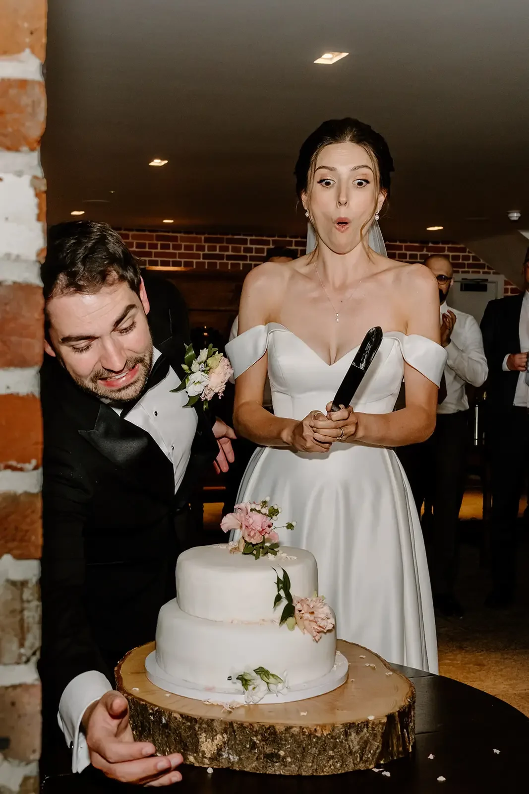 Bride and groom cutting wedding cake, bride with surprised expression holding a cake knife, groom smiling, decorated wedding cake with flowers on a wooden slab, indoor reception with brick walls, other guests in formal attire in background.
