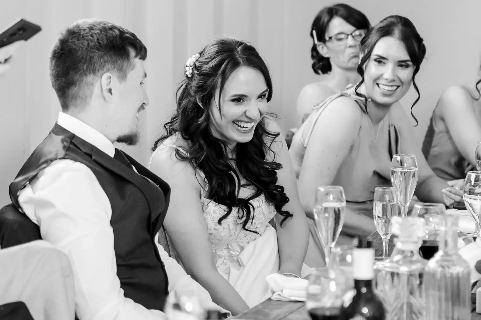 Black and white photo of a group of people, including a man and two women, sitting at a table during a wedding reception, smiling and laughing.