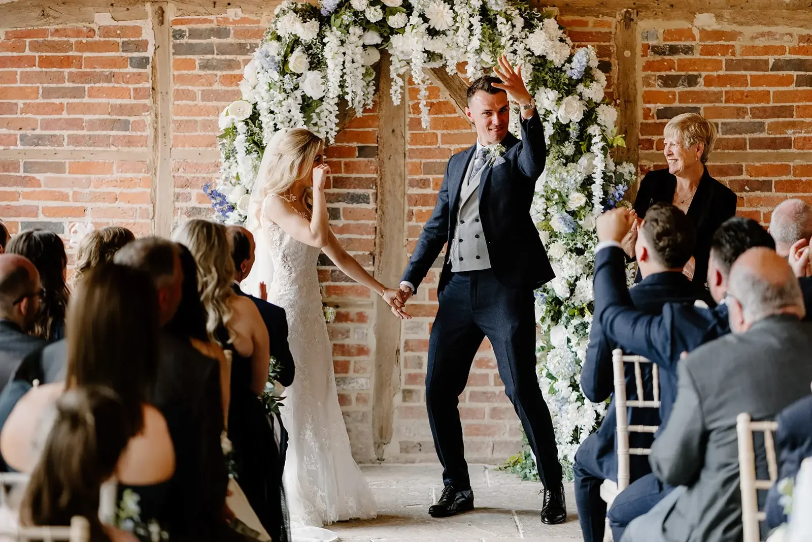 A bride and groom holding hands during their wedding ceremony with guests seated and an officiant standing nearby, in front of a floral arch against a brick wall.