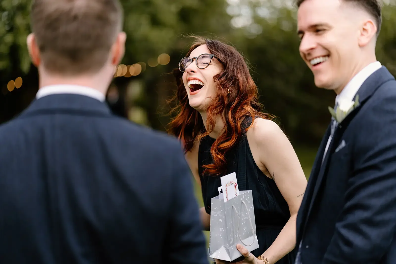 A woman with glasses and red hair laughing and holding a gift bag, standing outdoors with two men in suits.