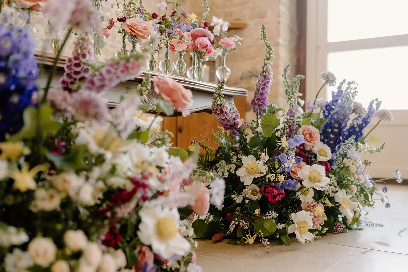 Various colorful flowers arranged on the floor and on a table near a window, with bottles of flowers on the table and a brick wall in the background.