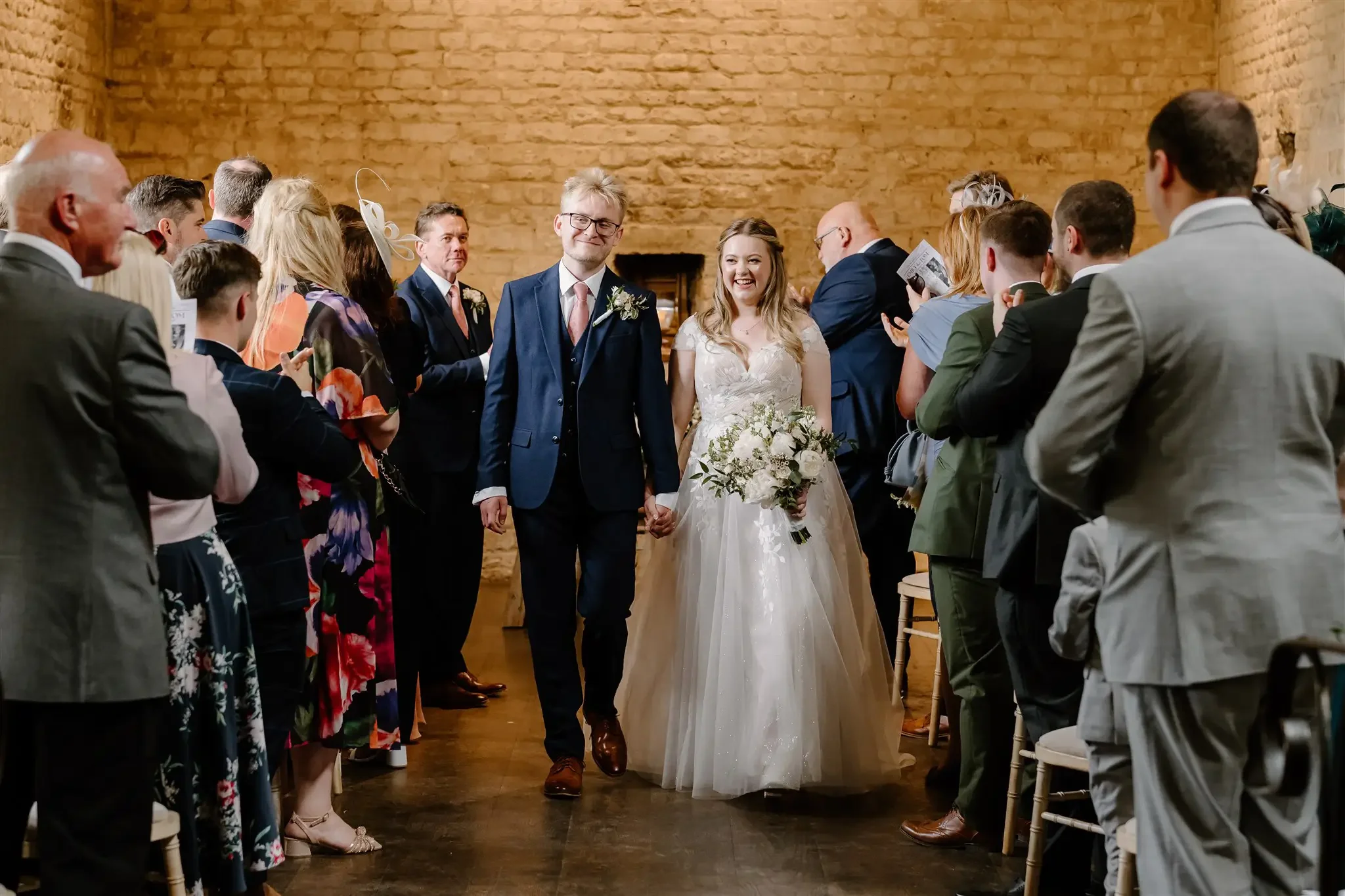 A bride and groom walking hand in hand down the aisle during their wedding ceremony, surrounded by seated guests in a warmly lit room with brick walls.
