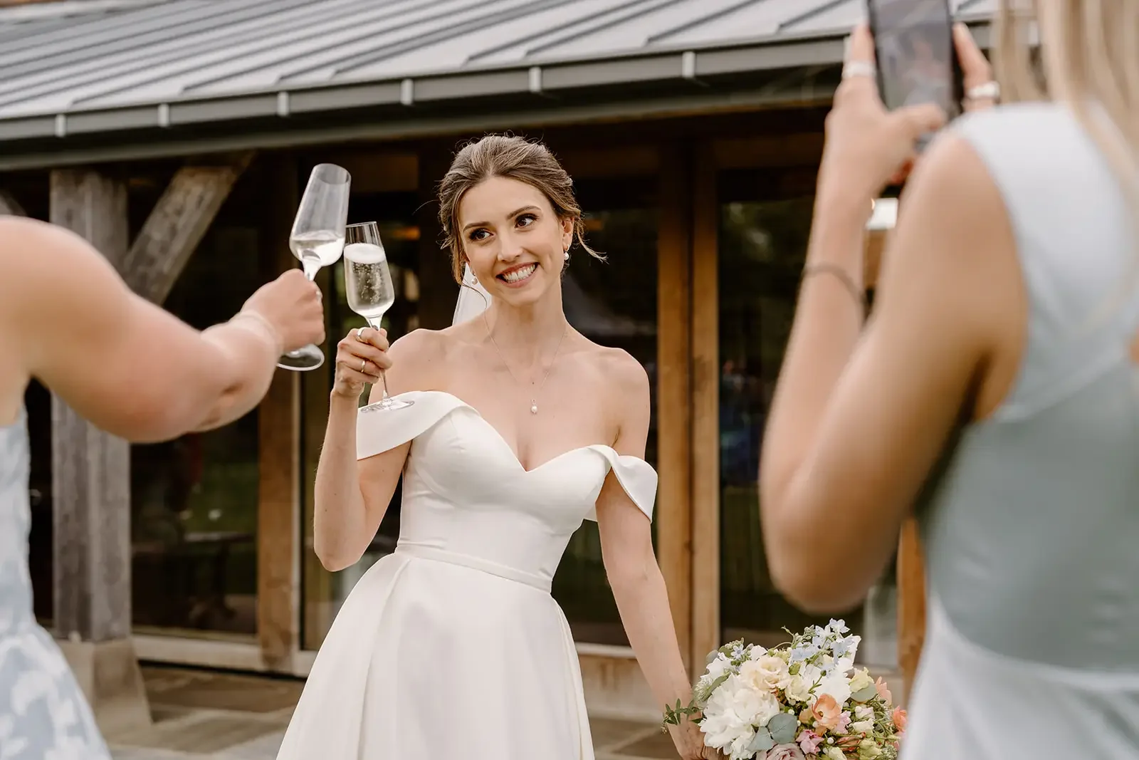 A bride in a white wedding dress smiling and holding a champagne glass, surrounded by friends at an outdoor celebration.