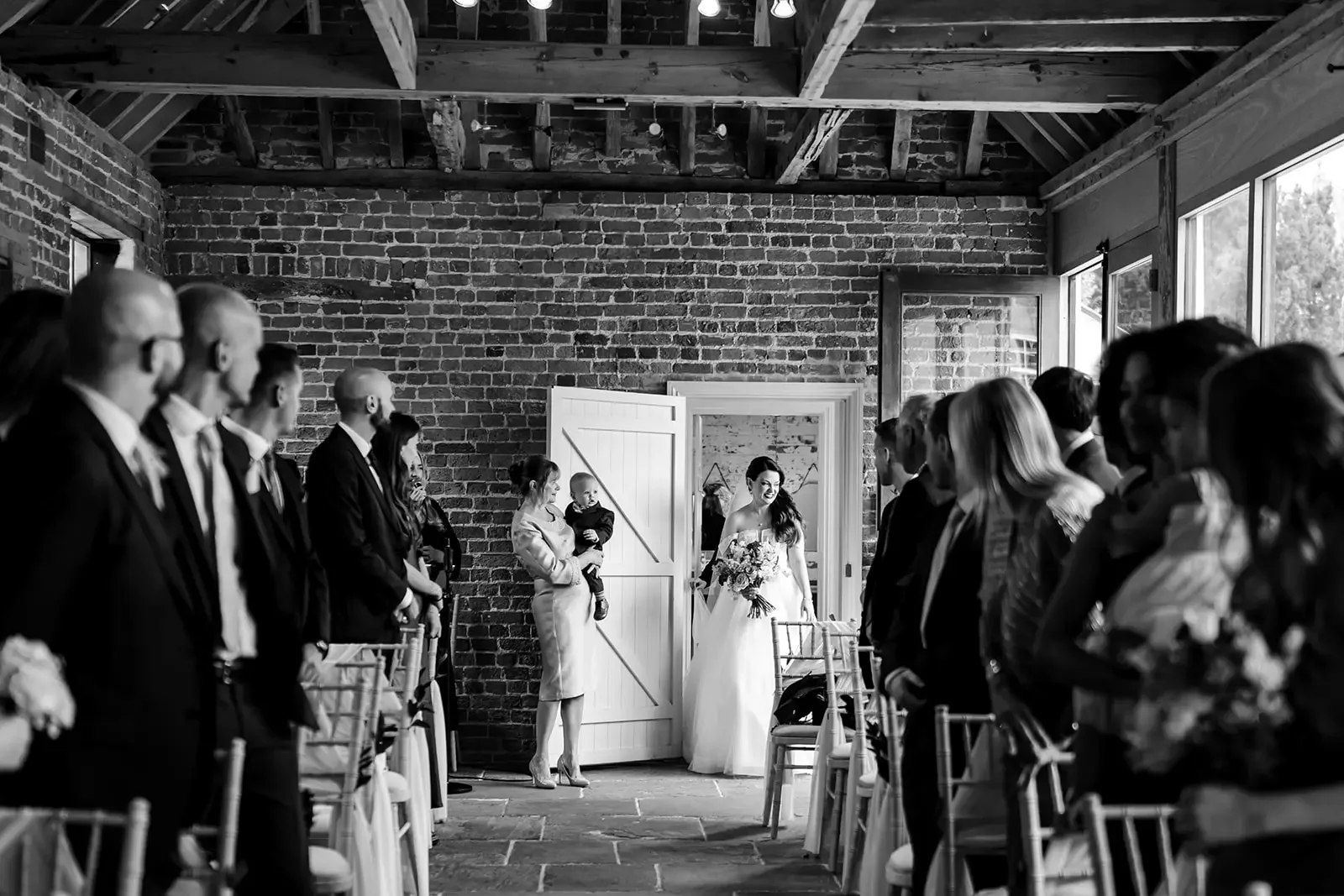 Bride walking down the aisle at her wedding ceremony in a rustic barn with brick walls, surrounded by seated guests.