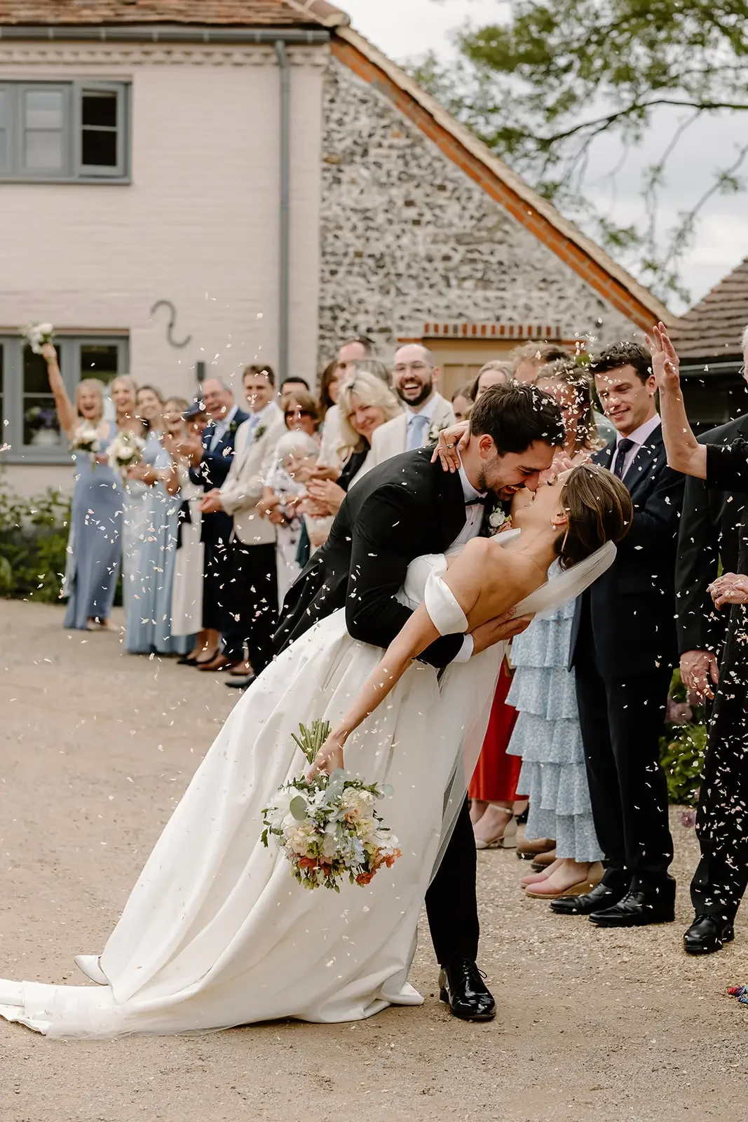 A newlywed couple kisses while their wedding party and guests celebrate and throw confetti outside a house.