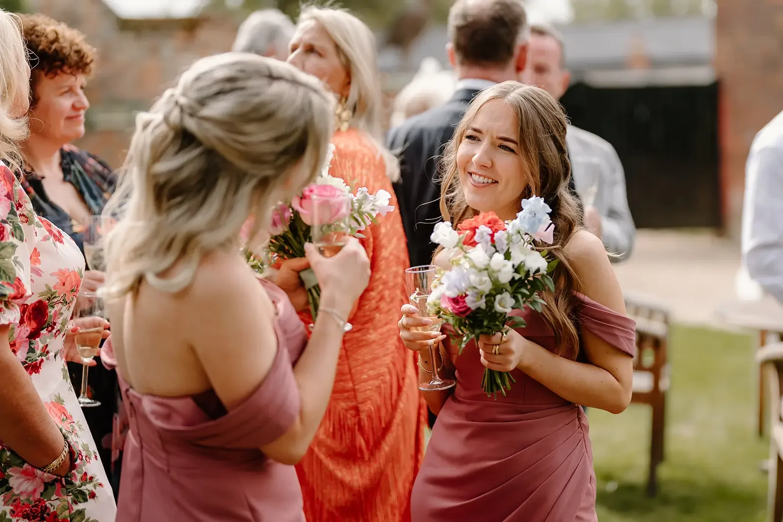 A group of women at a wedding reception outdoors, with one woman in a pink dress holding a bouquet of flowers and laughing, surrounded by other women in colorful dresses holding glasses, engaged in conversation.