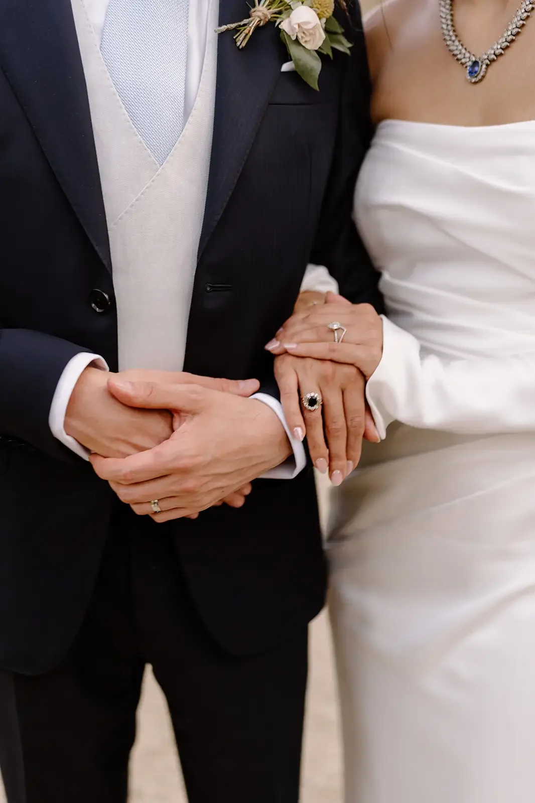 Close-up of a bride and groom holding hands during a wedding ceremony, showing wedding rings and formal attire.