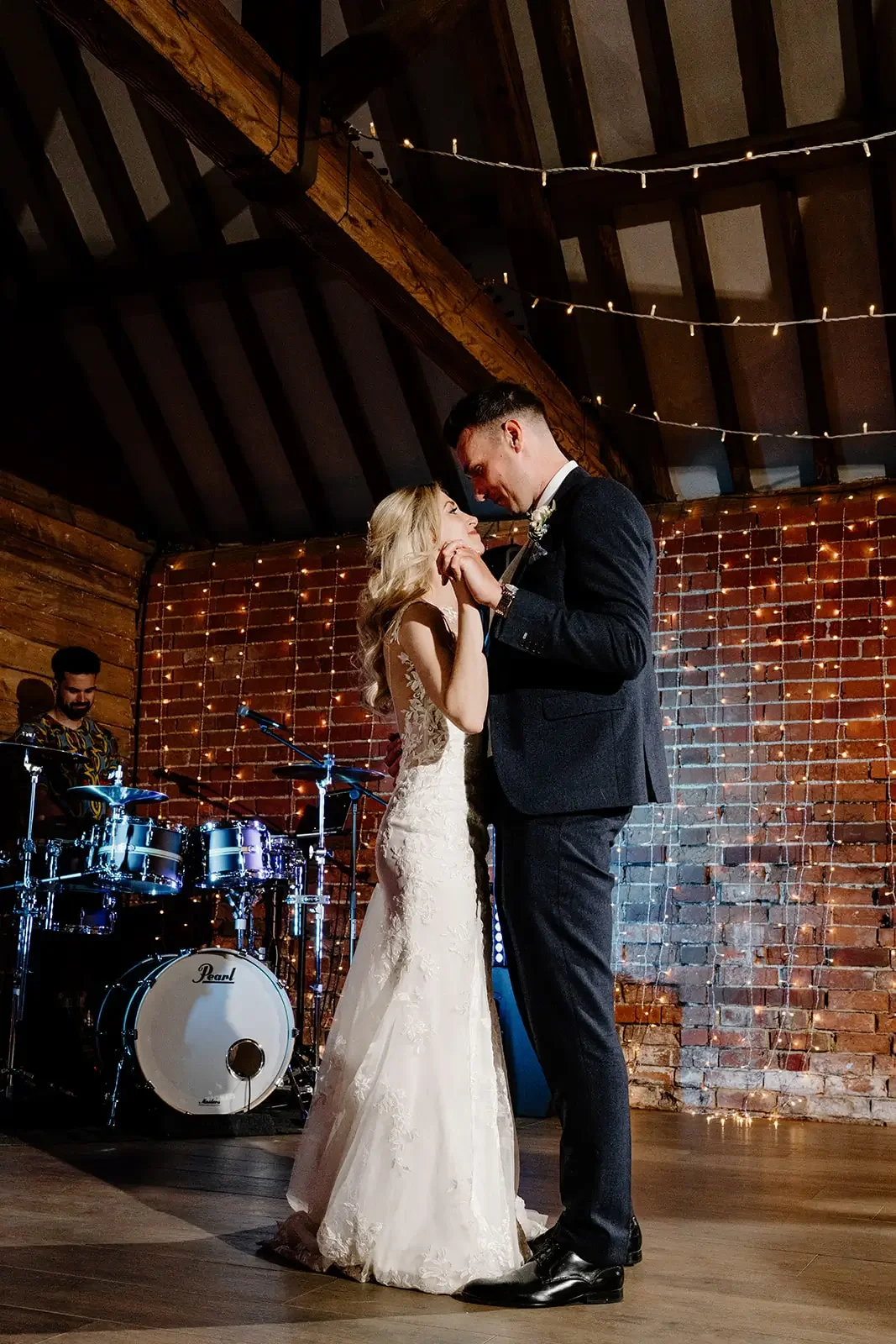 A bride and groom dancing at their wedding reception with a brick wall and string lights in the background, a drummer playing in the background.